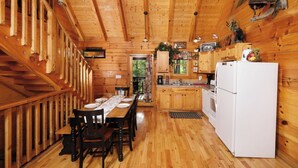 A wooden cabin interior with a dining table set for four, a kitchen with a refrigerator, and a staircase with wooden railings.