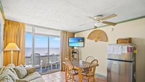 A room with a dining table, chairs, a sofa, a refrigerator, a television, and a view of the beach.