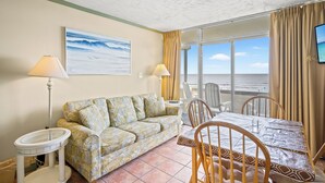 A room with a sofa, a dining table, chairs, a TV, and a view of the ocean.