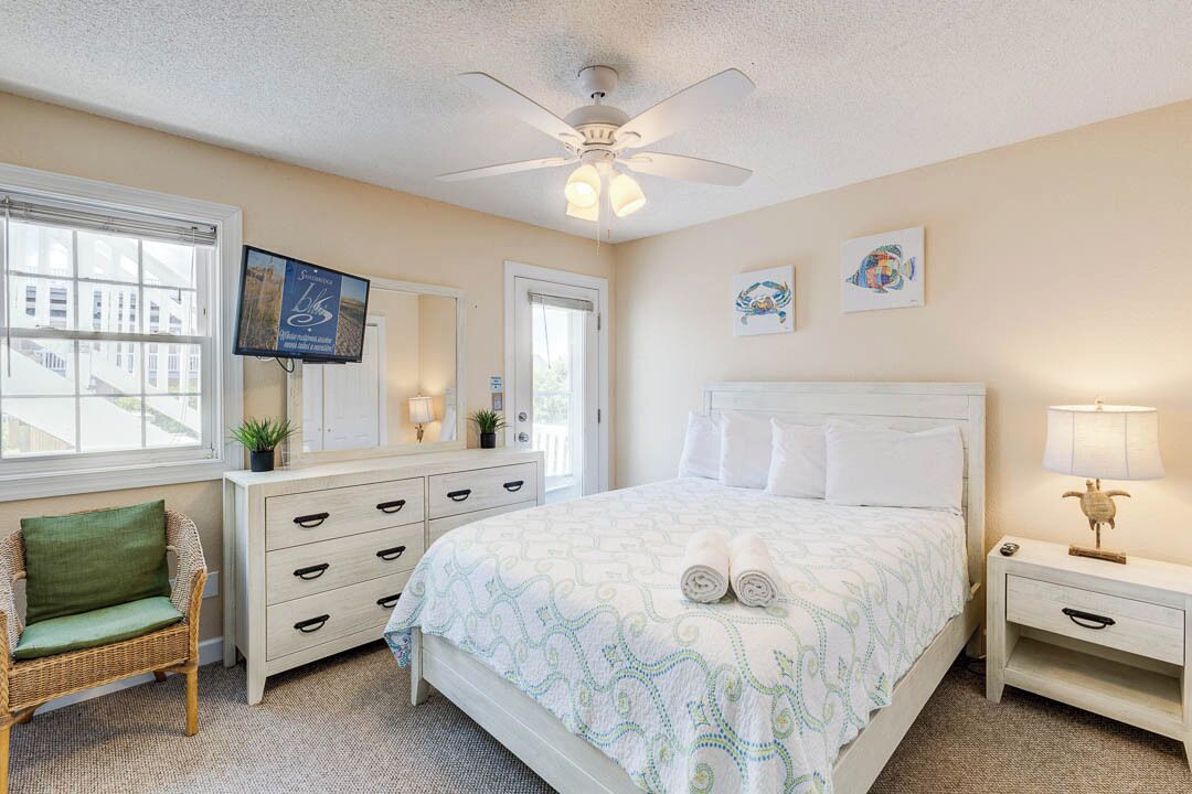 A bedroom with a bed, a dresser, a chair, a nightstand, a ceiling fan, and artwork on the walls.