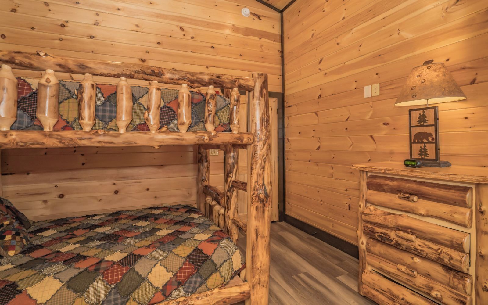 A rustic cabin room with a bunk bed, a wooden dresser, and a lamp.