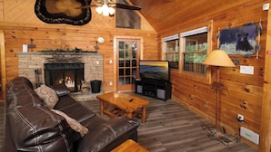 A cozy log cabin interior with a leather sofa, a fireplace, a television, and a bear-themed wall decoration. - Heaven on Earth - Pet Friendly & Secluded (Sevierville)