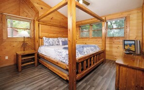 A wooden cabin bedroom with a four-poster bed, a television, and a window with a view of trees.