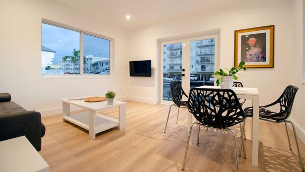 A modern living room with a large window, a flat-screen TV, a dining area with black chairs, and a painting on the wall. - Modern Apart beside canal, near beach w/parking (Sunny Isles Beach)
