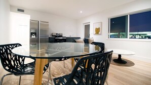 Un comedor moderno con una mesa de vidrio y sillas negras, una cocina al fondo y un ventanal con vistas a la ciudad.