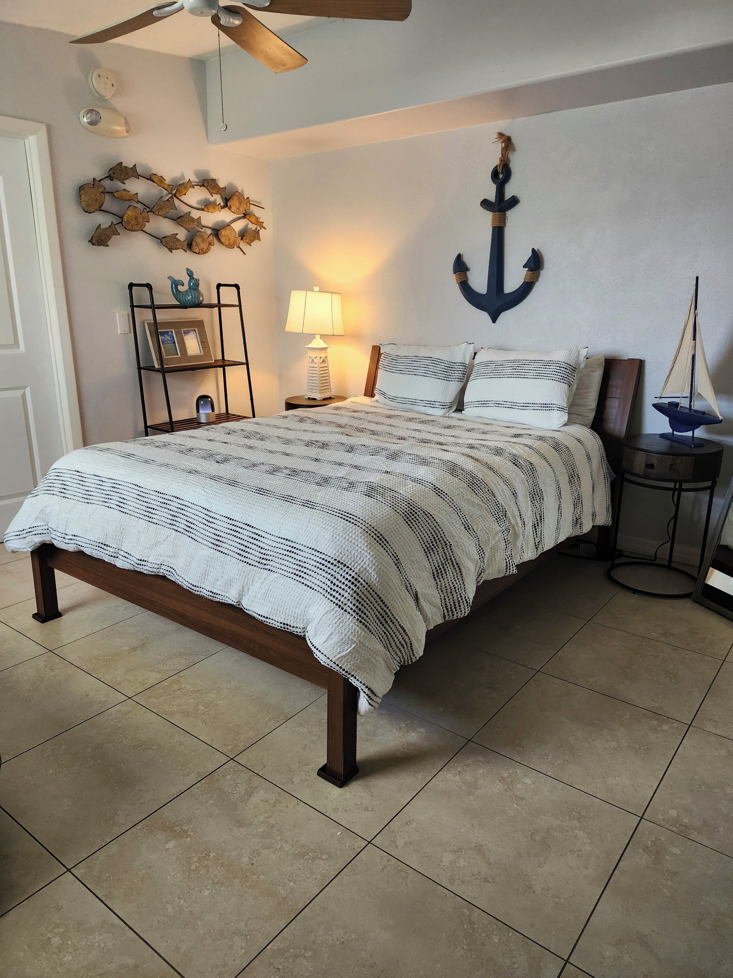 Una cama bien hecha con un edredón estampado, un adorno ancla en la pared, un pequeño estante con objetos decorativos y una lámpara sobre una mesita de noche.