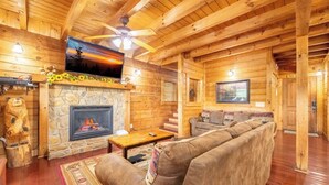 A cozy log cabin interior with a stone fireplace, a wooden ceiling, a sofa, and a television mounted above the fireplace. - Above It All - Pet Friendly, 3 Mi to Dollywood, Hot Tub + Pool Table & Games (Sevierville)