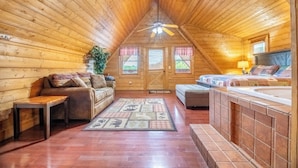A cozy attic bedroom with wooden walls, a ceiling fan, a sofa, a bed, and a hot tub. - Above It All - Pet Friendly, 3 Mi to Dollywood, Hot Tub + Pool Table & Games (Sevierville)