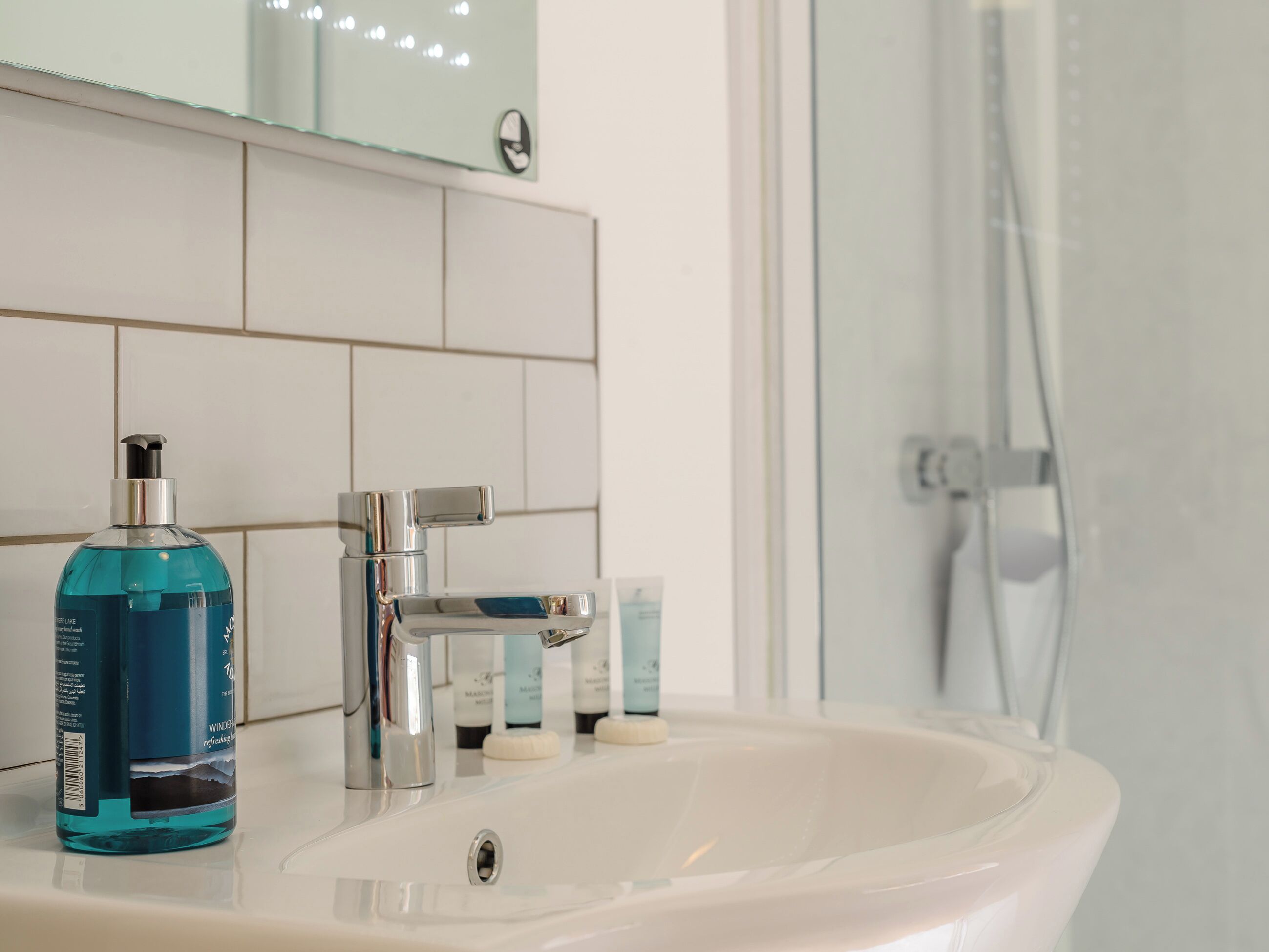 Een badkamer met een witte wastafel, een chromen kraan, een blauwe handzeepfles en een glazen douchecabine.