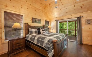 A wooden cabin bedroom with a bed, two bedside tables, a window with blinds, a balcony door, and a bear-themed wall decoration.