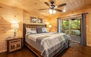 A wooden cabin bedroom with a bed, two bedside tables, a ceiling fan, and a view through a glass door.