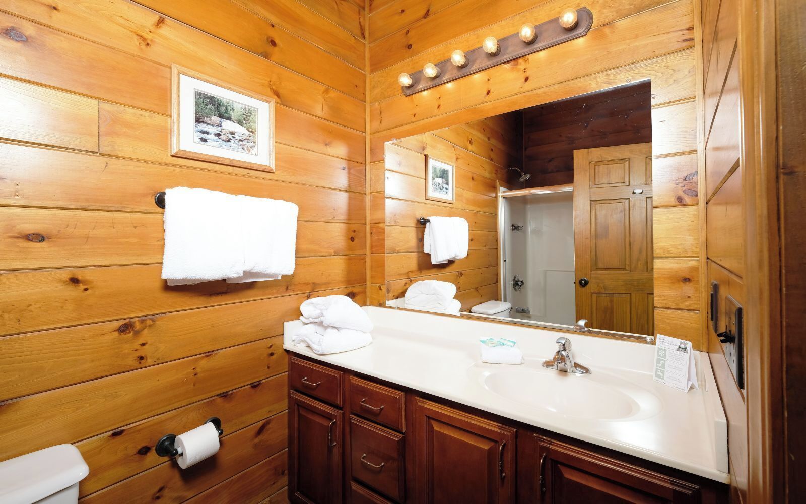 Een houten badkamer in chaletstijl, met een grote spiegel, houten kastjes en een witte wastafel.