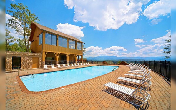 A pool area with lounge chairs, a stone wall, and a building with large windows. - Shooting Star - Stunning Views, Wi-Fi, Pool Table (Sevierville)