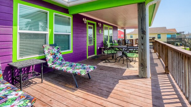 A covered outdoor deck with a purple and green exterior, a wooden floor, and a seating area with a colorful chair and table. - Seas The Moment before it passes you by. (Freeport)