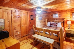Quarto de cabana de madeira com cama, um banco, um ventilador de teto e uma televisão.