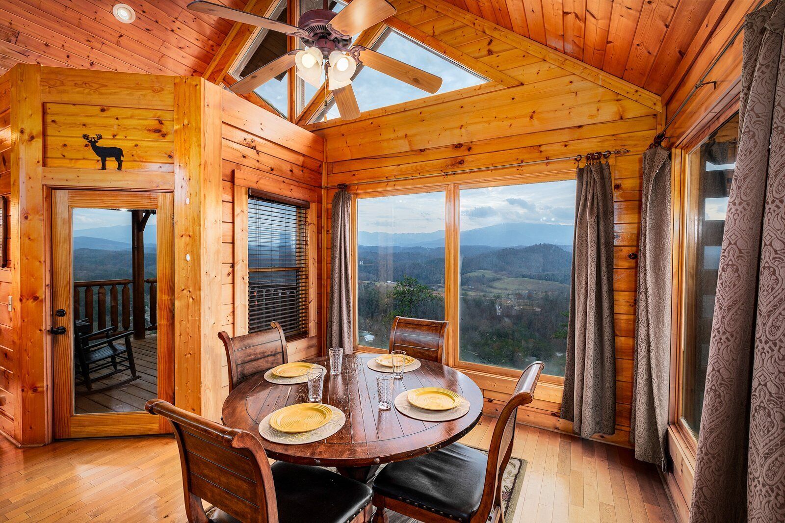 A wooden cabin with a dining table set for four, large windows offering a scenic view, and a ceiling fan.