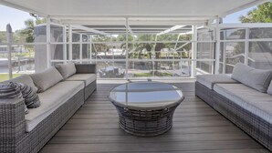 Een overdekt terras met rieten meubels en een tafel met glazen blad, met uitzicht op een jachthaven en palmbomen.