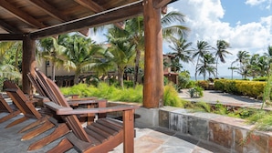 A covered outdoor seating area with wooden chairs and tables, surrounded by palm trees and a view of the ocean.