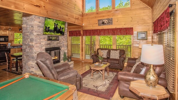 A cozy cabin interior with a pool table, leather sofas, a fireplace, and a television mounted on a stone wall. - 🐻Can't Bear to Leave: Theater, Two King Suites, Games, Fireplaces & Hot Tub🌄 (Pigeon Forge)