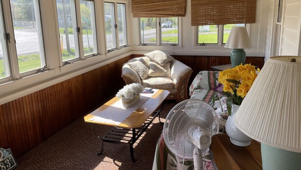 A cozy room with a wooden ceiling, a cushioned chair, a table with a fan, and a view of a road and greenery outside.
