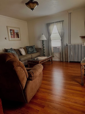 A living room with a brown sofa, a wooden coffee table, a lamp, a radiator, and a window with curtains.