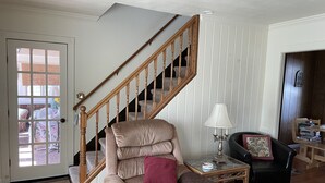 A living room with a beige armchair, a wooden staircase, a small table with a lamp, a black leather chair, and a bookshelf.