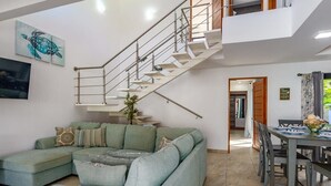A modern living room with a green sectional sofa, a dining area with a wooden table, and a staircase with a metal railing.