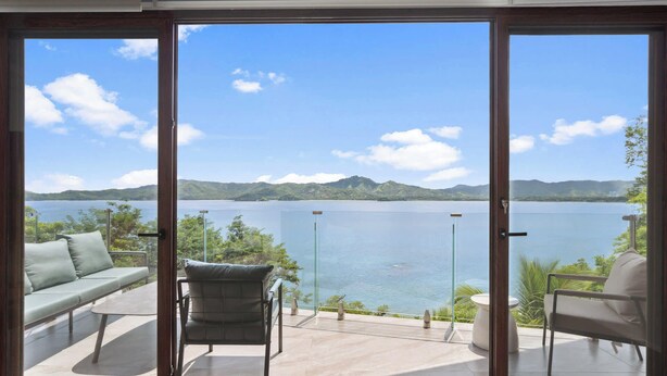 A balcony with a glass door, a sofa, a chair, and a table, overlooking a lake with mountains in the distance. - 5-BD Beachfront, Ocean-View Hillside Home (Playa Flamingo)