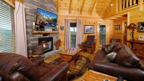 A cozy log cabin interior with a stone fireplace, leather sofas, and a television mounted on the wall. - Still Life - Mountain Views and Hot Tub (Sevierville)