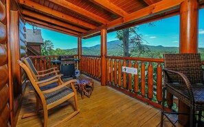 A covered porch with wooden chairs, a grill, and a view of a mountainous landscape.