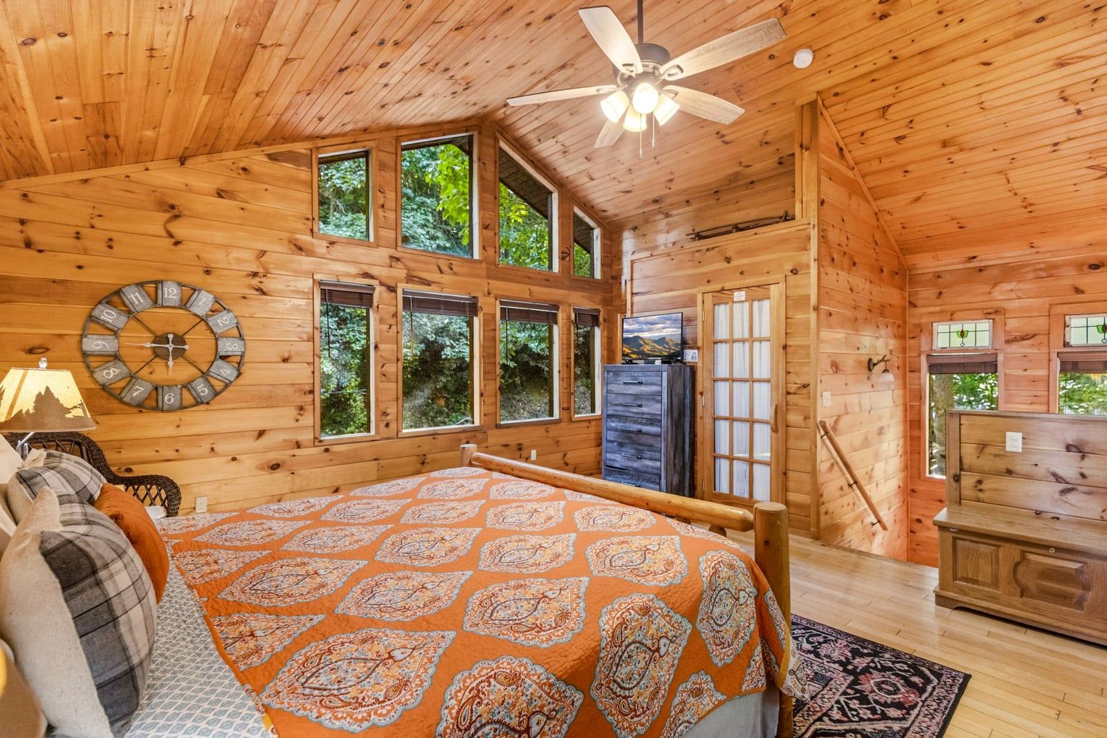 A wooden cabin bedroom with a bed, a clock, a TV, and large windows.