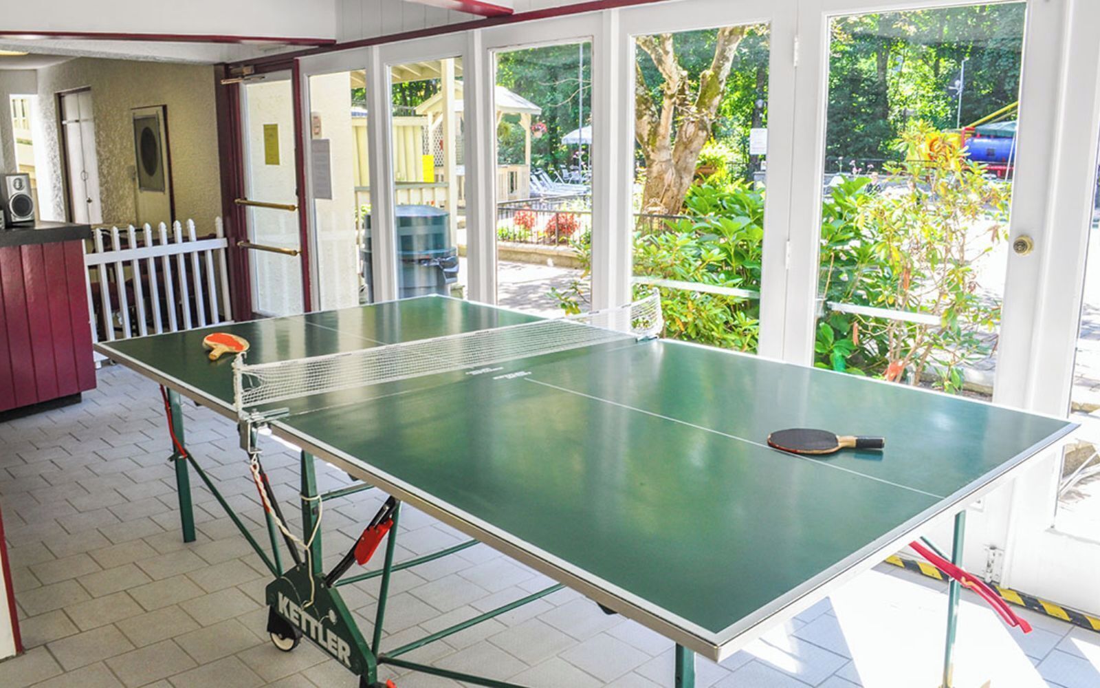 Una mesa de ping pong con red, una raqueta y una base Kettler en una habitación con ventanas amplias y vistas a zonas verdes.