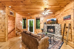 A cabin interior with a stone fireplace, wooden walls, a sofa, a coffee table, and a ceiling fan. - Smoky Getaway - Hot tub hangs and cozy nights! (Gatlinburg)