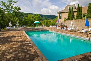 Piscina con sillas de descanso y sombrillas, rodeada por un patio de ladrillo y un muro de piedra.