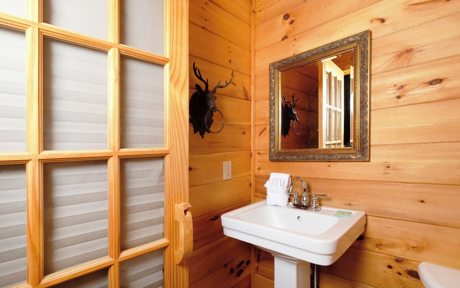 A wooden cabin bathroom with a wooden-framed window, a sink with a towel, and a deer head mounted on the wall.
