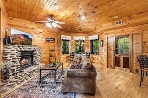 Ein Interieur einer Holzhütte mit Kamin aus Stein, Holzwänden, Deckenventilator und Essbereich.