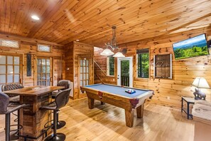 Interior de una cabaña de madera con mesa de billar, comedor y televisor montado en la pared.