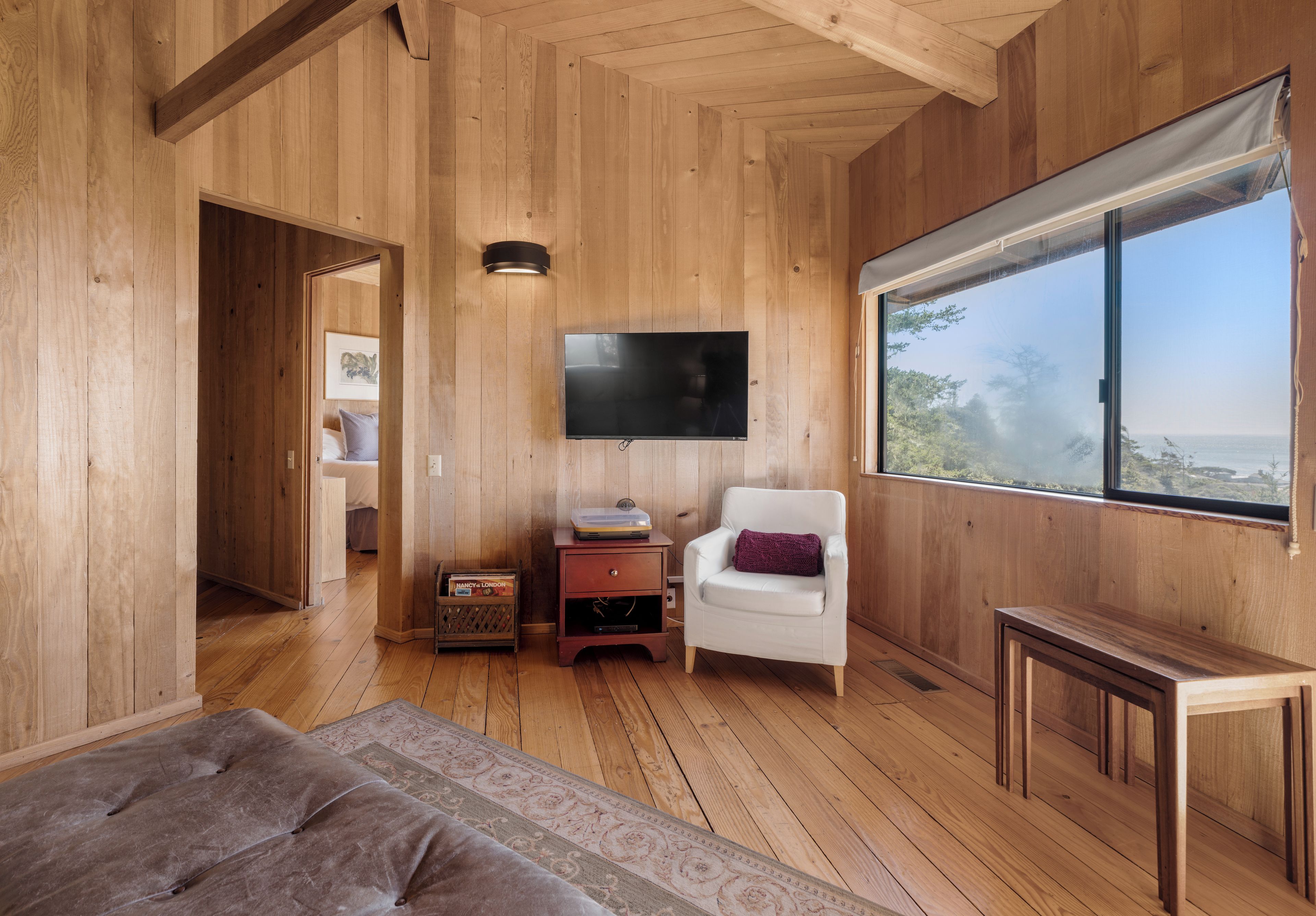 Een houten interieur met een flatscreen televisie aan de muur, een witte fauteuil met paars kussen, en een groot raam met uitzicht op de omgeving.
