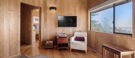 Een houten interieur met een flatscreen televisie aan de muur, een witte fauteuil met paars kussen, en een groot raam met uitzicht op de omgeving.
