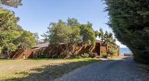 A wooden cabin with a gravel driveway, surrounded by trees and a clear view of the ocean.