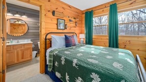 A wooden cabin bedroom with a bed, a bathroom with a sink and mirror, and a window with curtains. - Mountain VIEWS! Hot Tub, Handicap Accessible - 15 mins to downtown Gatlinburg (Sevierville)