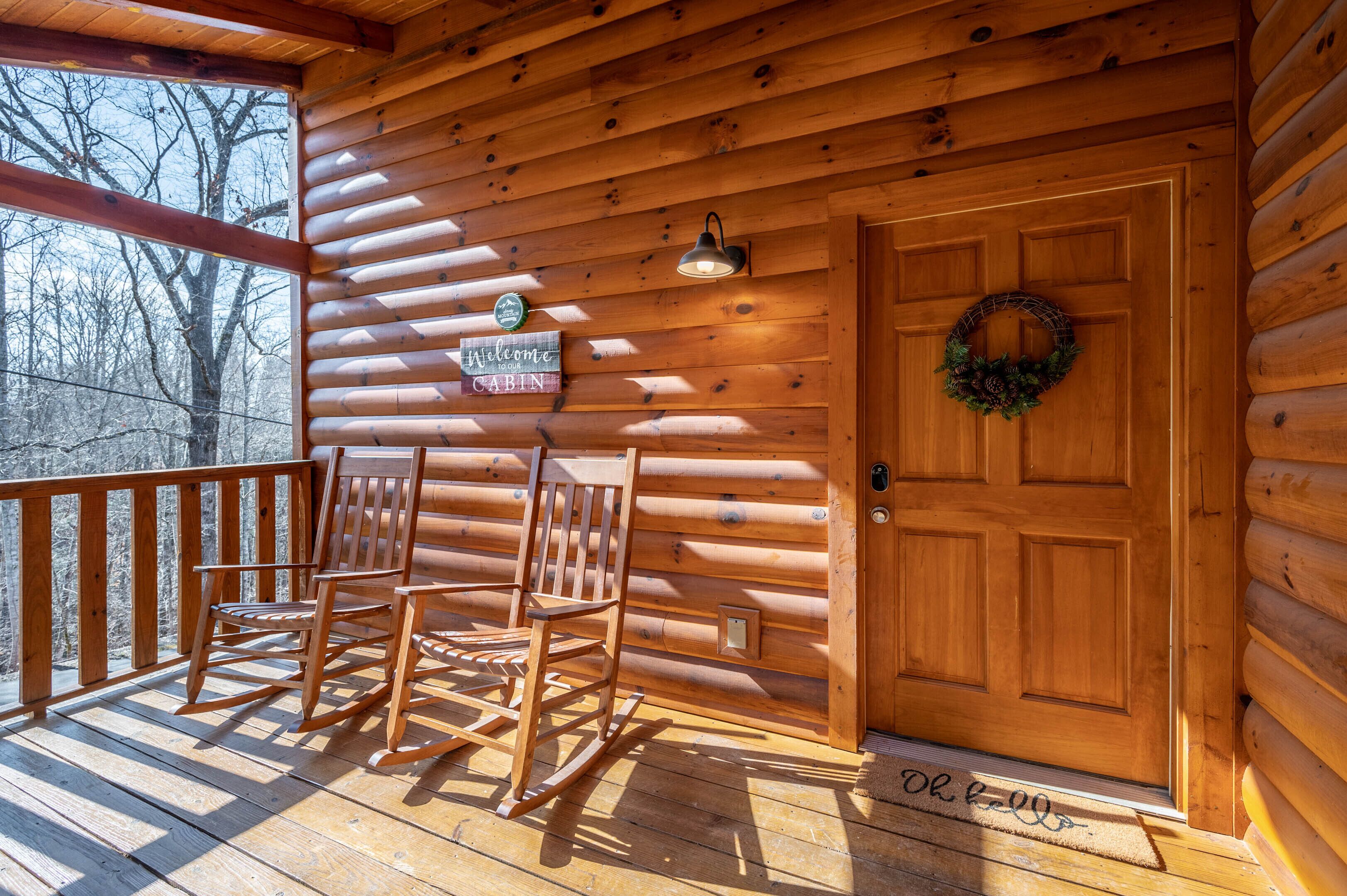 A wooden cabin with a porch, rocking chairs, a door with a wreath, and a welcome sign.