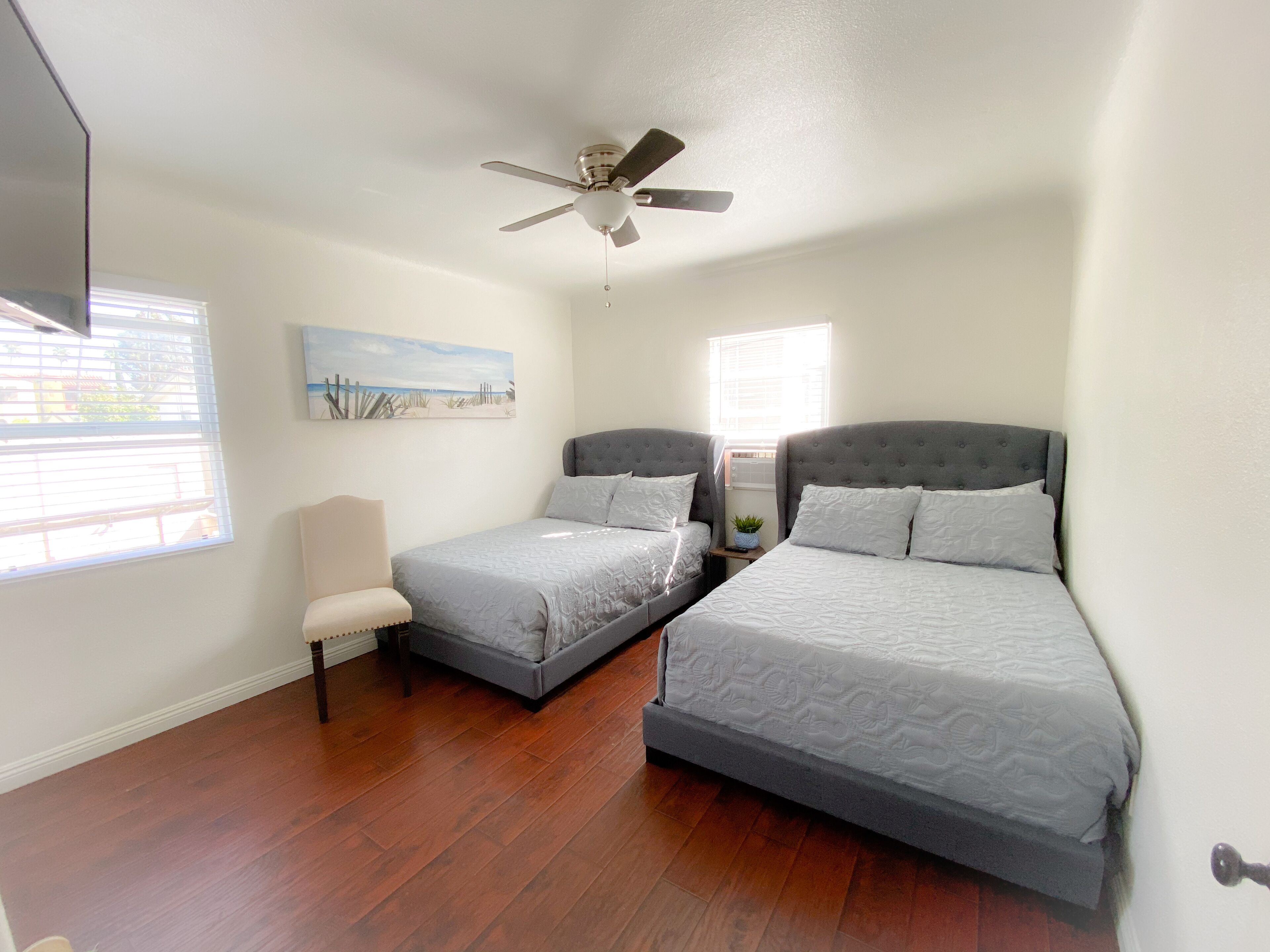 A bedroom with two beds, a ceiling fan, a painting on the wall, and a window with blinds.