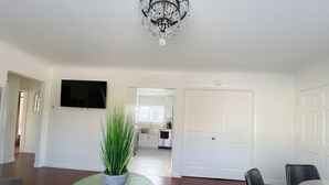A living room with a round glass coffee table, a potted plant, a chandelier, and a TV mounted on the wall.