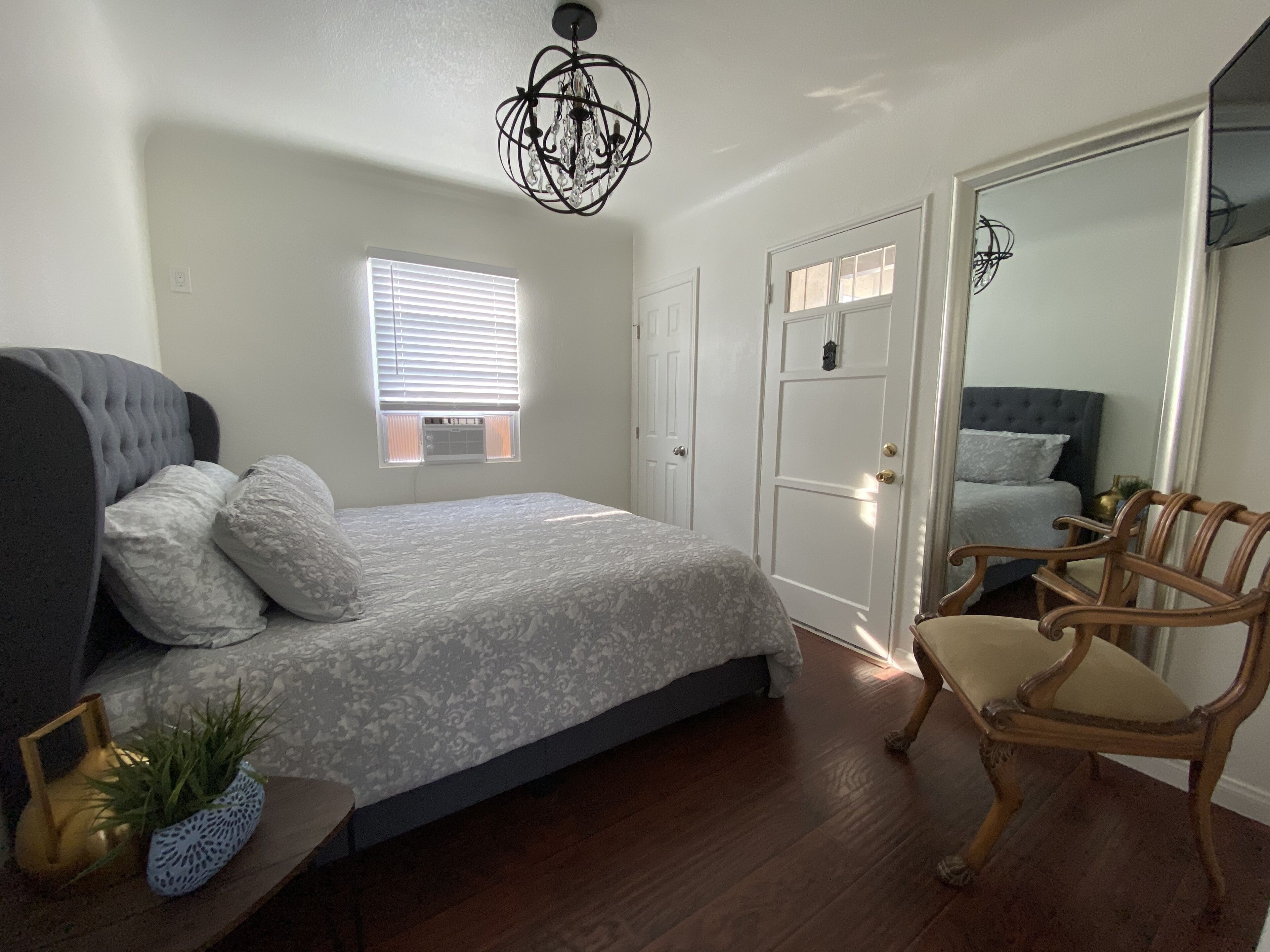 Une chambre à coucher comprenant un lit, une chaise, une table de chevet avec une plante, une fenêtre avec des stores et une porte.