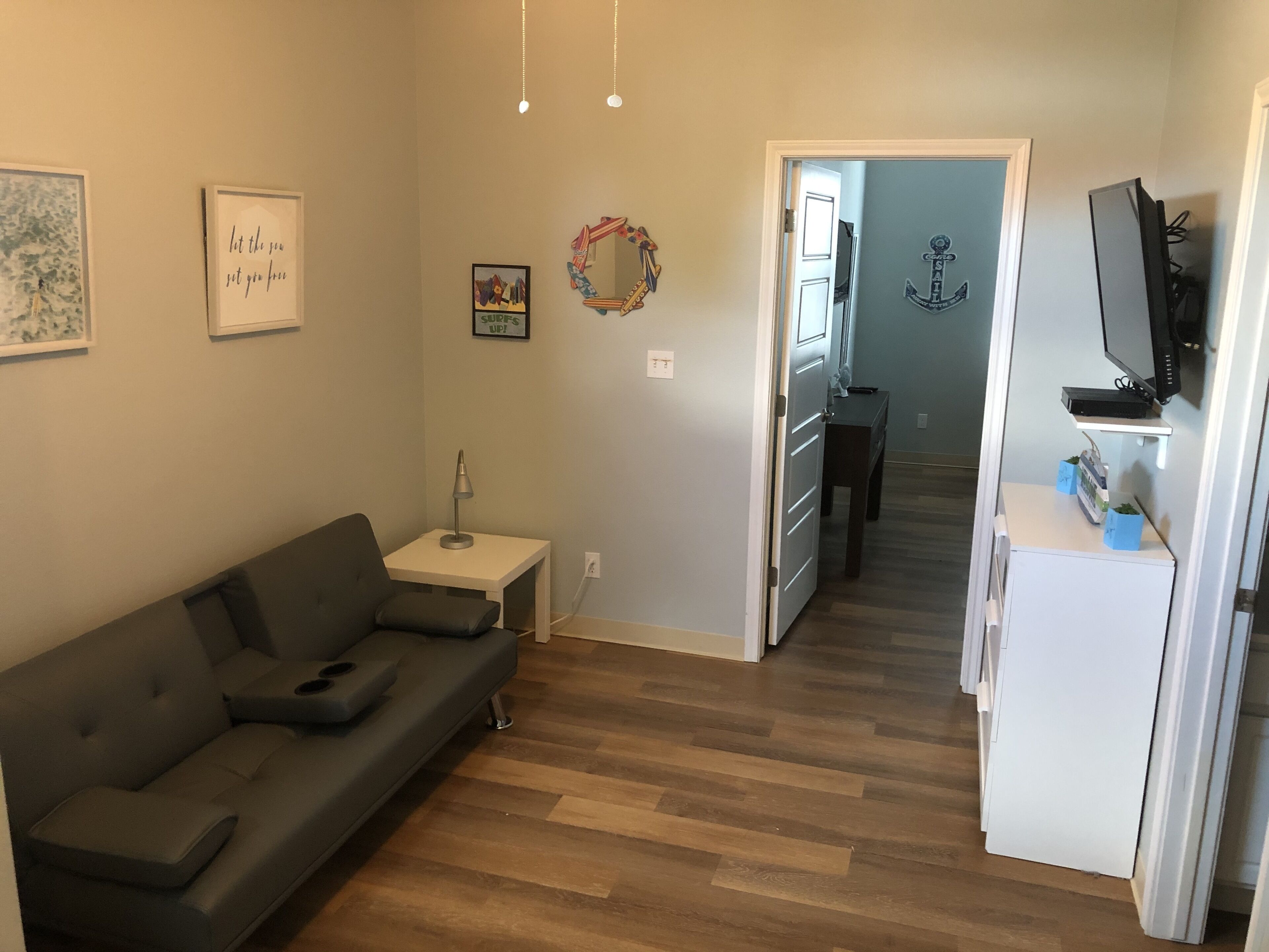 A living room with a gray sofa, a small side table with a lamp, and a kitchen area with a refrigerator and a TV.