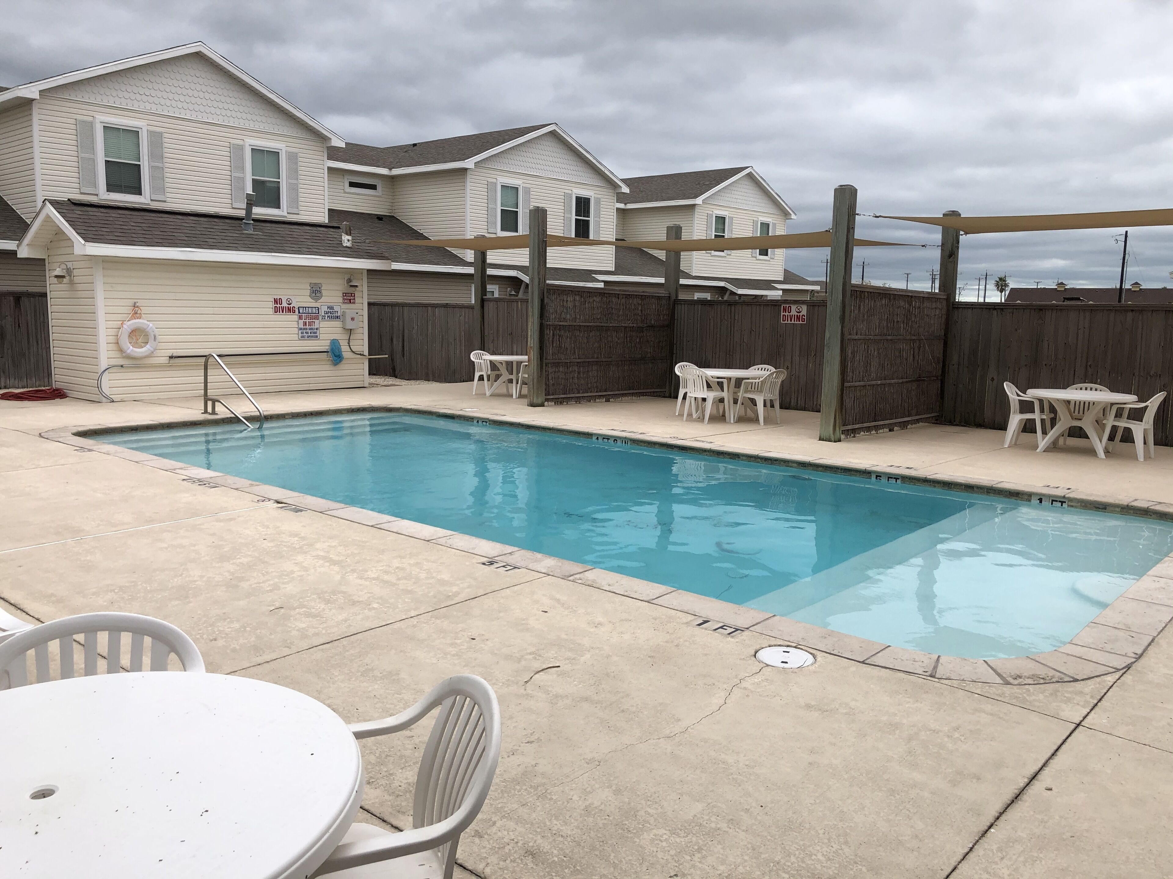Une piscine rectangulaire avec une terrasse en bois et un panneau indiquant « Interdiction de plonger ». On y trouve des chaises et une table en plastique, toutes deux de couleur blanche, disposées autour de la piscine.
