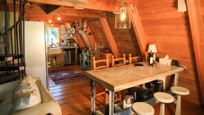 Un interior rústico de madera con comedor que incluye una mesa y taburetes, cocina con ollas colgantes y una zona de estar acogedora con sofá y sillón.