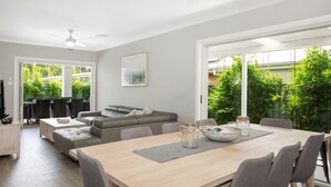 A modern living room with a dining area, grey sofas, a wooden dining table, and a view of greenery outside. - Callala Bay Beach House - Wowly Waters - Pet Friendly 🐳 (Callala Bay)