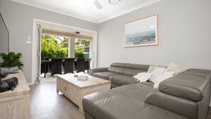 A modern living room with a sectional sofa, a coffee table, and a dining area with chairs and a table. - Callala Bay Beach House - Wowly Waters - Pet Friendly 🐳 (Callala Bay)
